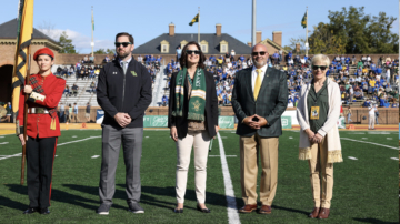 Five people stand on a football field. One wears a military uniform and holds a flag