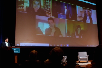 Drew Stelljes stands at a podium on stage near a large screen showing several people