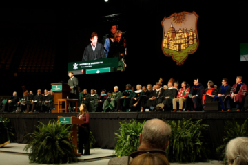 People in academic regalia sit on a stage while people stand at podiums