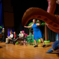 Several people sit in chairs on a stage while watching a person stand and gesture next to an inflatable octopus.
