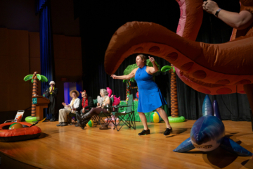 Several people sit in chairs on a stage while watching a person stand and gesture next to an inflatable octopus.