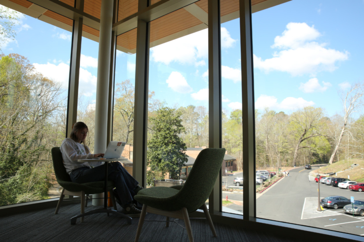 A William & Mary student works on a computer while sitting next to a large window.