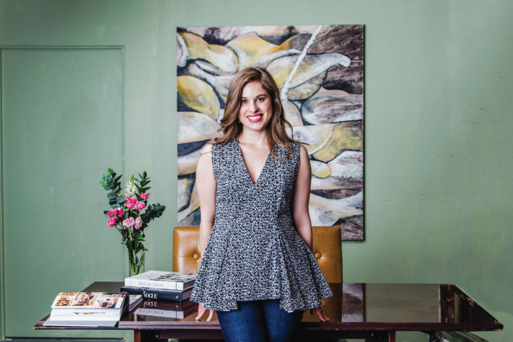 Delia Folk stands in front of a desk with a large piece of artwork behind it.