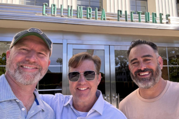 Three people pose in front of a building with a Columbia Pictures sign.