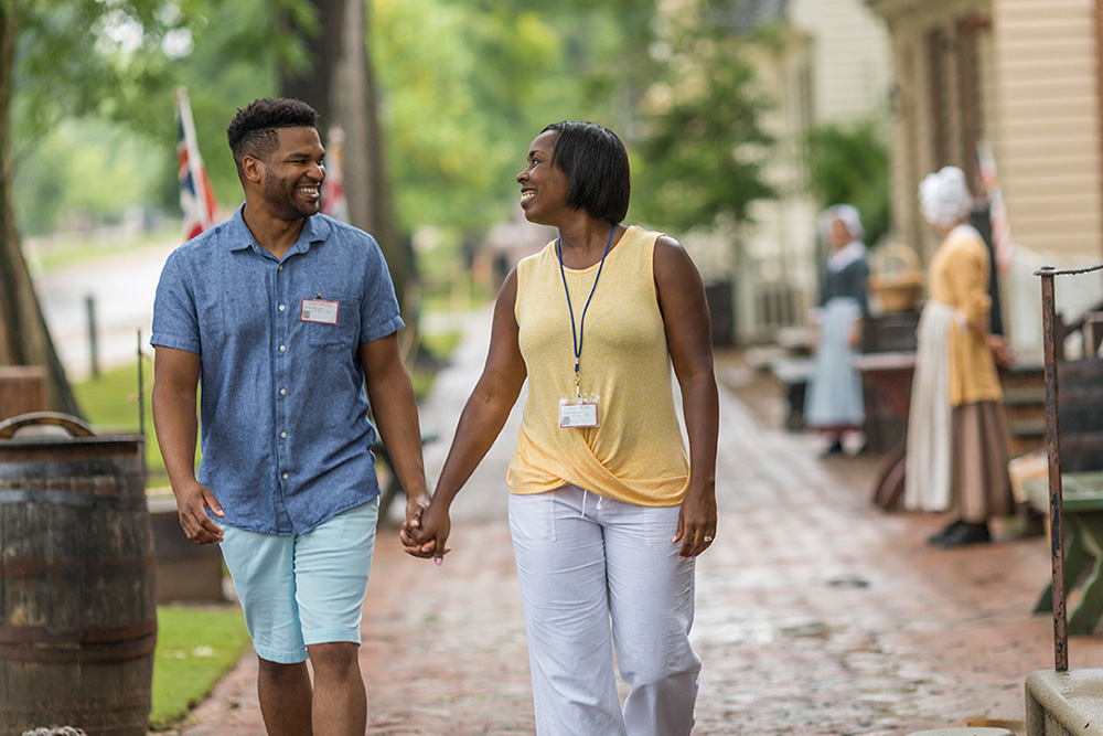 A smiling couple walking hand in hand down a brick sidewalk with Colonial Williamsburg interpreters in the background