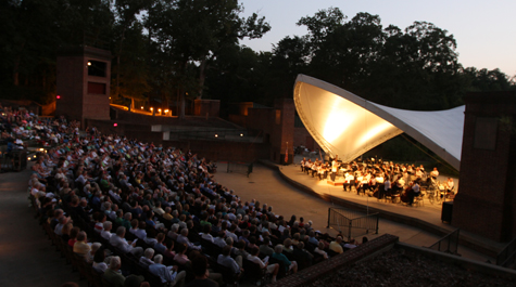 People sit in an outdoor concert venue facing a stage with a cover