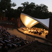 People sit in an outdoor concert venue facing a stage with a cover