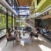 Students sit and work on computers in a lobby surrounded by glass windows