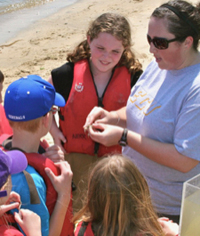 Children wearing life jackets surround an adult on a beach