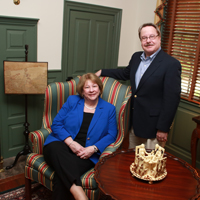 One person sits in a chair and another person stands by the chair inside a living room