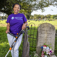 A person stands next to a headstone and holds a weedwhacker