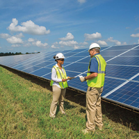 Two people in hard hats stand near solar panels in a large field
