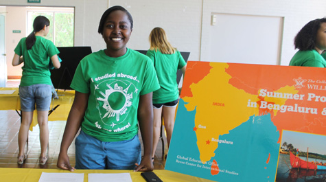 A person wearing a shirt saying "I studied abroad" stands behind a table with a poster showing India on it