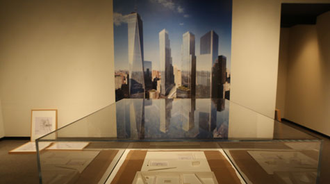 A large photo of the World Trade Center memorial on the wall behind a glass case containing flat white architectural models related to the site.