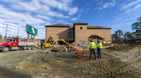 Two people in construction hats stand in a construction area outside of a brick building
