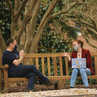 Two people wearing masks talk while sitting on a bench