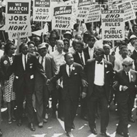 A black and white photo shows a large group of people marching and holding signs about employment