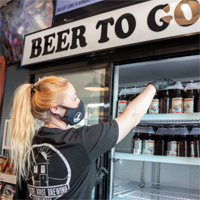 A person wearing a mask and gloves reaches into a cooling case that says Beer To Go on the top