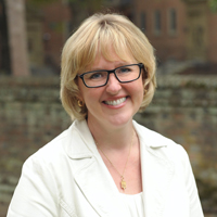 Ginger Ambler in a white suit stands against a brick wall