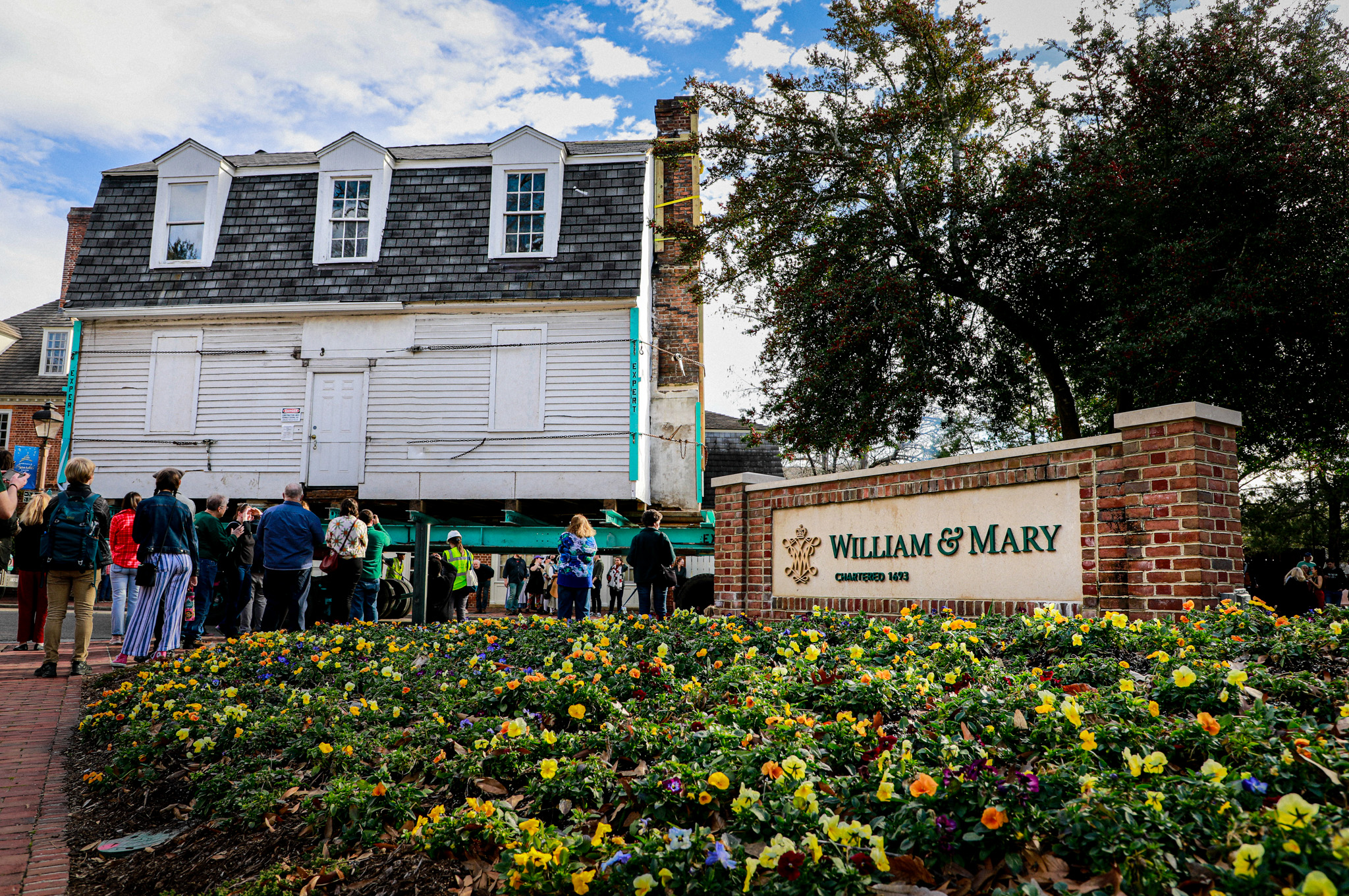 Behind a William & 玛丽 sign, the Bray School Lab building is physically moved with a trailer across campus, with onlookers watching from the side.